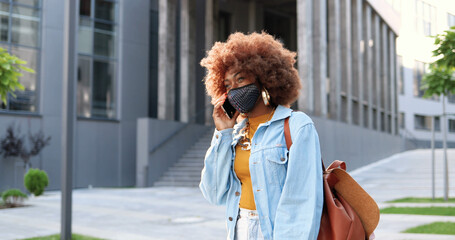 Fototapeta premium African American young stylish pretty woman with curly hair wig and in mask standing at street, talking on telephone. Female outside in city speaking on mobile phone. Pandemic or pollution concept.