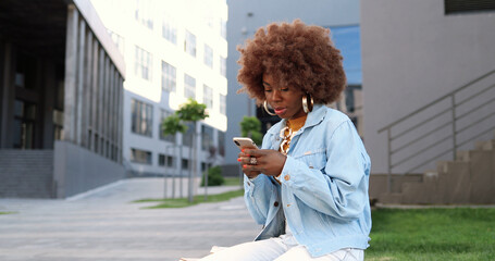 African American beautiful woman tapping and scrolling on mobile phone and sitting on bench outside. Stylish attractive female messaging on smartphone. Telephone chatting.