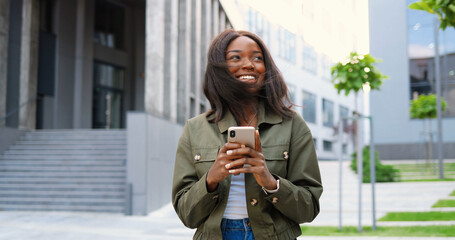 African American joyful young stylish woman tapping or scrolling on smartphone and standing at city street. Beautiful happy female texting message on mobile phone and smiling. Outside.