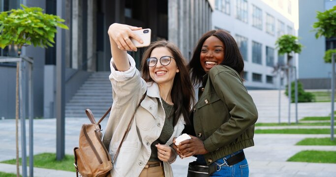 Mixed-races young pretty females smiling cheerfully to camera and taking selfie photo with mobile phone camera at city street.