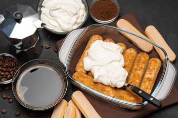 tiramisu dessert cooking - Traditional Italian Savoiardi ladyfingers Biscuits and cream in glass baking dish, coffe maker on dark concrete background