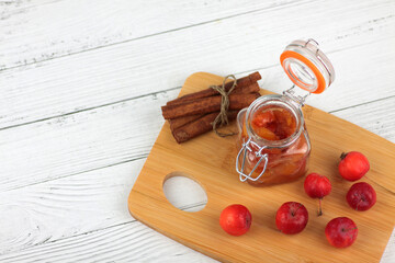 glass jar of apple jam, red apples and cinnamon sticks on cutting board on white wooden background flat lay