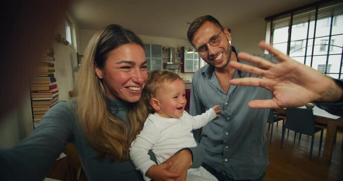 A happy smiling family with toddler baby boy is making a selfie or video call to friends or relatives while having a breakfast together in a living room at home.