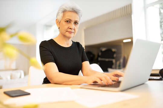 Indoor image of attractive confident middle aged female copywriter working remotely from home typing article for online magazine, sitting at desk in front of open portable computer, looking at camera