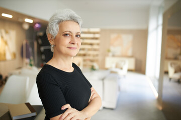 Indoor image of happy attractive retired European female with short gray hair standing in stylish living room keeping arms crossed on chest, her posture expressing confidence. Age and lifestyle