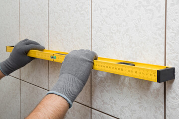 Worker hands in protective gloves measuring tiles surface on wall with yellow spirit level....
