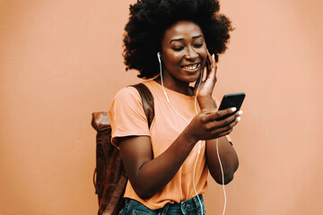 Young smiling african hipster woman having earphones on and listening music over smart phone while standing outdoors.