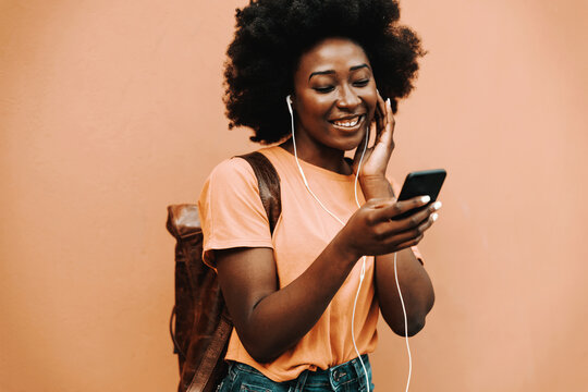 Young smiling african hipster woman having earphones on and listening music over smart phone while standing outdoors.