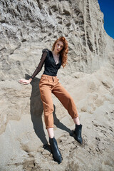 Fashion red-Haired girl in orange jeans posing in nature near sandy rocks, not like everyone else