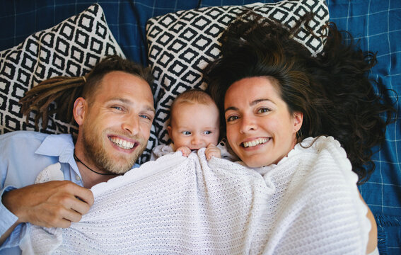 Young couple with baby daughter resting under blanket, looking at camera.