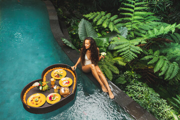 Girl relaxing and eating floating breakfast in jungle pool on luxury villa in Bali. Valentines day or honeymoon surprise. Tropical travel lifestyle. Black rattan tray in heart shape.