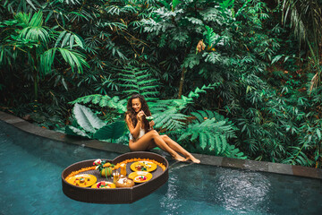 Girl relaxing and eating floating breakfast in jungle pool on luxury villa in Bali. Valentines day or honeymoon surprise. Tropical travel lifestyle. Black rattan tray in heart shape. - 382989084