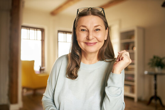 Portrait of happy cheerful European woman pensioner with eyeglasses on her head relaxing at home, sitting in stylish living room, looking at camera with joyful smile. People, age and lifestyle concept