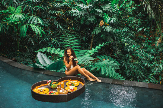 Girl relaxing and eating floating breakfast in jungle pool on luxury villa in Bali. Valentines day or honeymoon surprise. Tropical travel lifestyle. Black rattan tray in heart shape.
