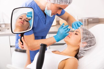 Female client looking in mirror during procedure of face mesotherapy in professional clinic of...