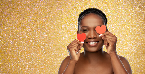 beauty, valentine's day and luxury concept - portrait of happy smiling young african american woman with bare shoulders with red hearts covering her eye over golden glitter background