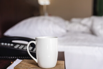 White cup of hot coffee on night table with unmade bed in background.