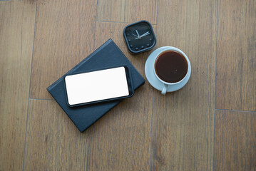 top view of blank screen smartphone on black book and cup of coffee on wooden background.