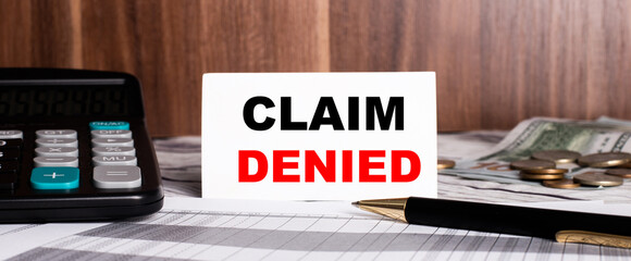 CLAIM DENIED written on a white card on a wooden background near a calculator and a pen