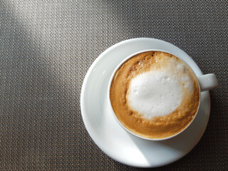 Cappuccino coffee with white and brown milk foam in a white cup and saucer stands on a table covered with a brown tablecloth.