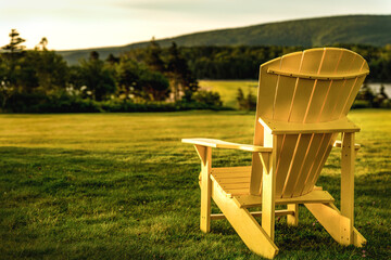 Canadian yellow adirondack chair on a grassy meadow, overlooking hazy hills at sunrise. Peaceful countryside scenery. Muskoka or Laurentian chair. Canada