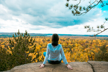 Young woman traveller sitting on a steep rock, admiring panoramic view of autumn forest in Algonquin Provincial Park, Ontario, Canada. Solo travelling, rear view