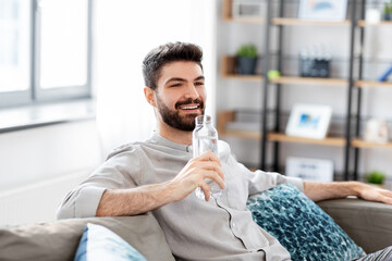 people, sustainability and leisure concept - happy smiling young man sitting on sofa and drinking water from glass bottle at home