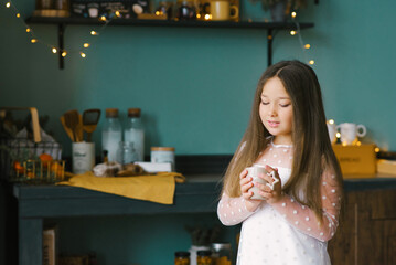 A beautiful girl warms her hands while holding a Cup of hot tea or cocoa among the Christmas decorations and looks at the Cup while standing in the kitchen