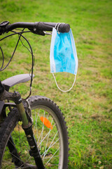 Fototapeta na wymiar A blue medical Mask hangs on handlebar of Bicycle against background of green Grass.
