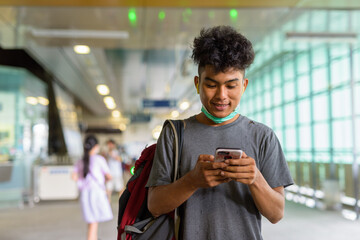 Happy young Asian tourist man as backpacker with mask using phone at the sky train station
