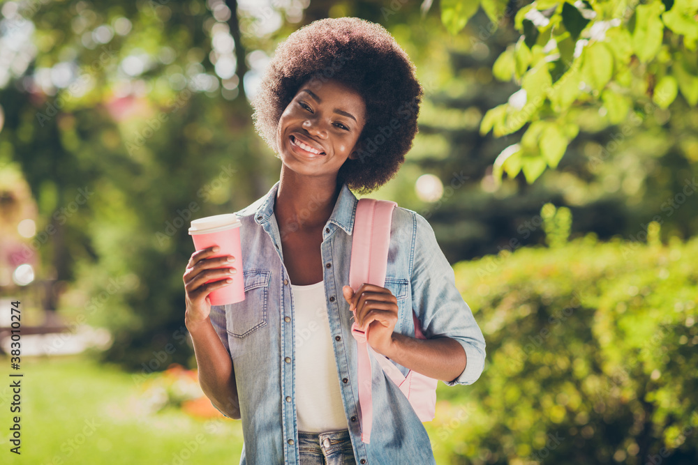 Poster Photo portrait of pretty cheerful black skinned curly girl keeping mug of hot drink walking in summer park wearing denim clothes backpack