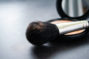 Close-up of a black makeup brush with flesh-colored powder at the end. On the background of an open box with a mirror and pink powder. Dark, almost black background. Copy space