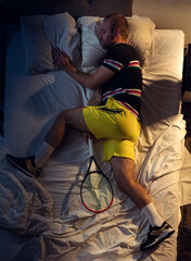 Champion. Top view of young professional tennis player sleeping at his bedroom in sportwear with racket. Loving his sport even more than comfort, watching match even if resting. Action, motion, humor.