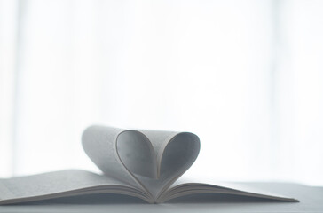 Twisted pages as hear sign. love concept. Soft focus Love story book with open page of literature in heart shape on the white table next to the window. Heart from a book page against the day light.