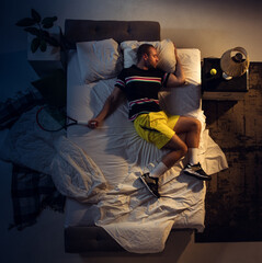 Fototapeta premium Champion. Top view of young professional tennis player sleeping at his bedroom in sportwear with racket. Loving his sport even more than comfort, watching match even if resting. Action, motion, humor.
