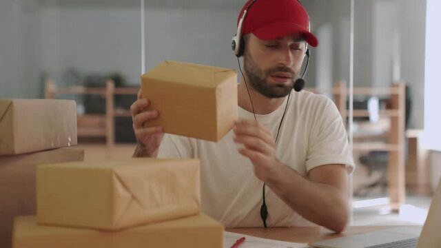 Young tired nervous post manager indoors in office using laptop computer and holding parcel boxes