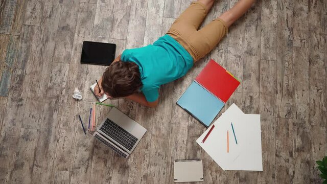 Top view of a boy surfing internet or learning online, lying on the wooden floor and using laptop, doing his homework while spending time alone at home