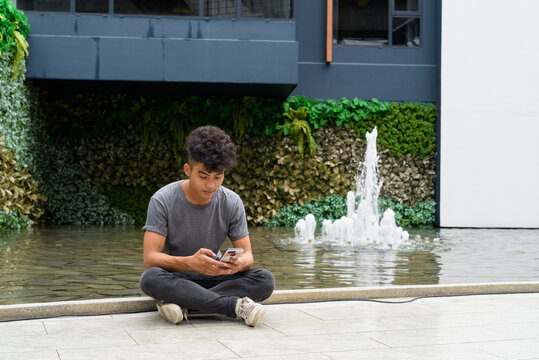 Young Asian Man Sitting While Using Phone Outside Modern Building