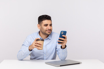 Elegant happy smiling businessman sitting with coffee in hand and checking message, reading email on mobile phone, taking break resting at workplace. indoor studio shot isolated on white background