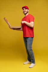 Photo of happy man from delivery service in red t-shirt and cap giving food order and holding pizza box isolated over yellow background