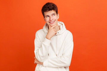 Smiling young man with peaceful face thinking over plans for future holding his chin, thoughtful teenager dreaming about good job. Indoor studio shot isolated on orange background