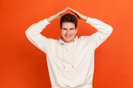 Happy smiling man in casual style sweater holding hands above head showing roof, housing on credit for youth, social security. Indoor studio shot isolated on orange background