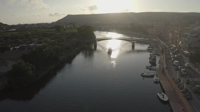 Aerial shot for a speed boat passing under a city bridge on Temo river in Sardinia island during sunset. Boats, cars, city buildings on one riverside and green land with trees on the other one.