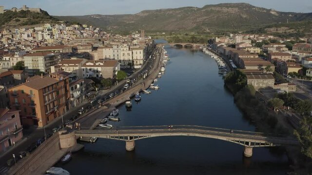Aerial push in shot for Temo river of Sardinia island with boats and colored buildings and few car traffic at the street. River bridges over the river canal and green mountains in the background