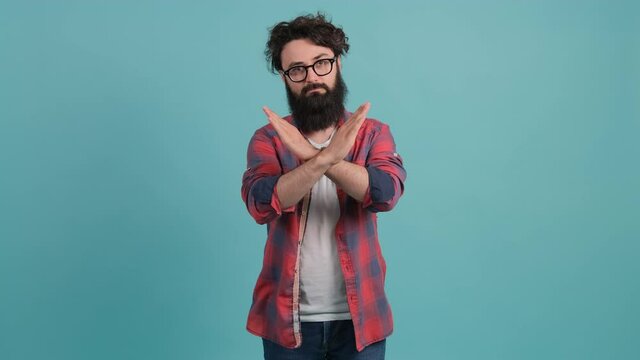 Portrait of a young bearded man annoyed with bad attitude making stop sign crossing hands, saying no, expressing security over turquoise background.