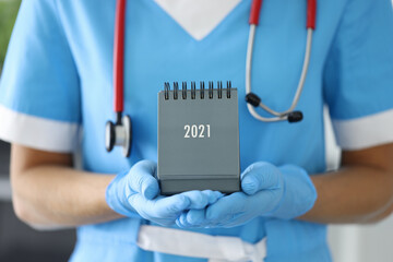 Doctor with phonendoscope around his neck holds desk calendar close-up in rubber gloves. Planning...