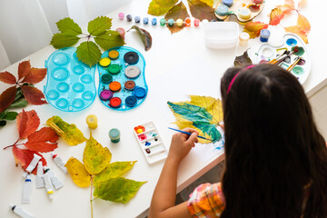 little girl paints leaves with brush at home, autumn art.