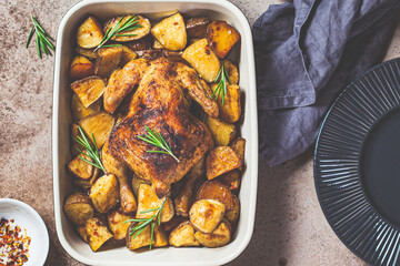 Baked whole  chicken with potatoes and rosemary in white dish, dark background, top view.