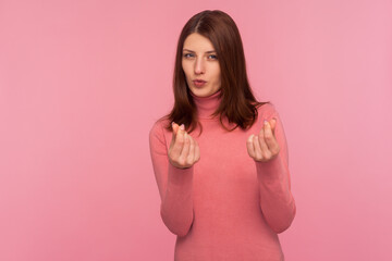 Greedy mercantile woman with brown hair in pink sweater asking cash showing money gesture with fingers seriously looking at camera. Indoor studio shot isolated on pink background