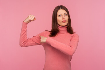 Strong self confident brunette woman in pink sweater showing her arm muscles raising hand, free and independent, emancipation. Indoor studio shot isolated on pink background